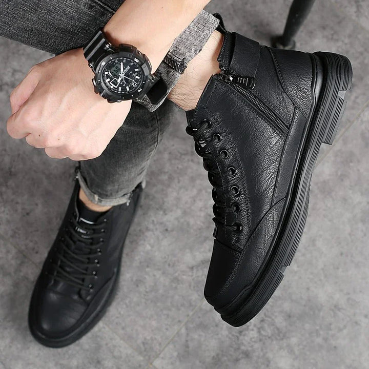 Stylish black high-top leather sneakers with zipper detail worn with rolled dark grey jeans and black wristwatch on grey floor
