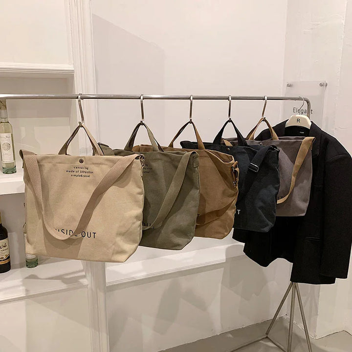 Row of five canvas tote bags in beige, green, brown, black, and gray hanging on a metal rack next to a black coat in a minimalist room