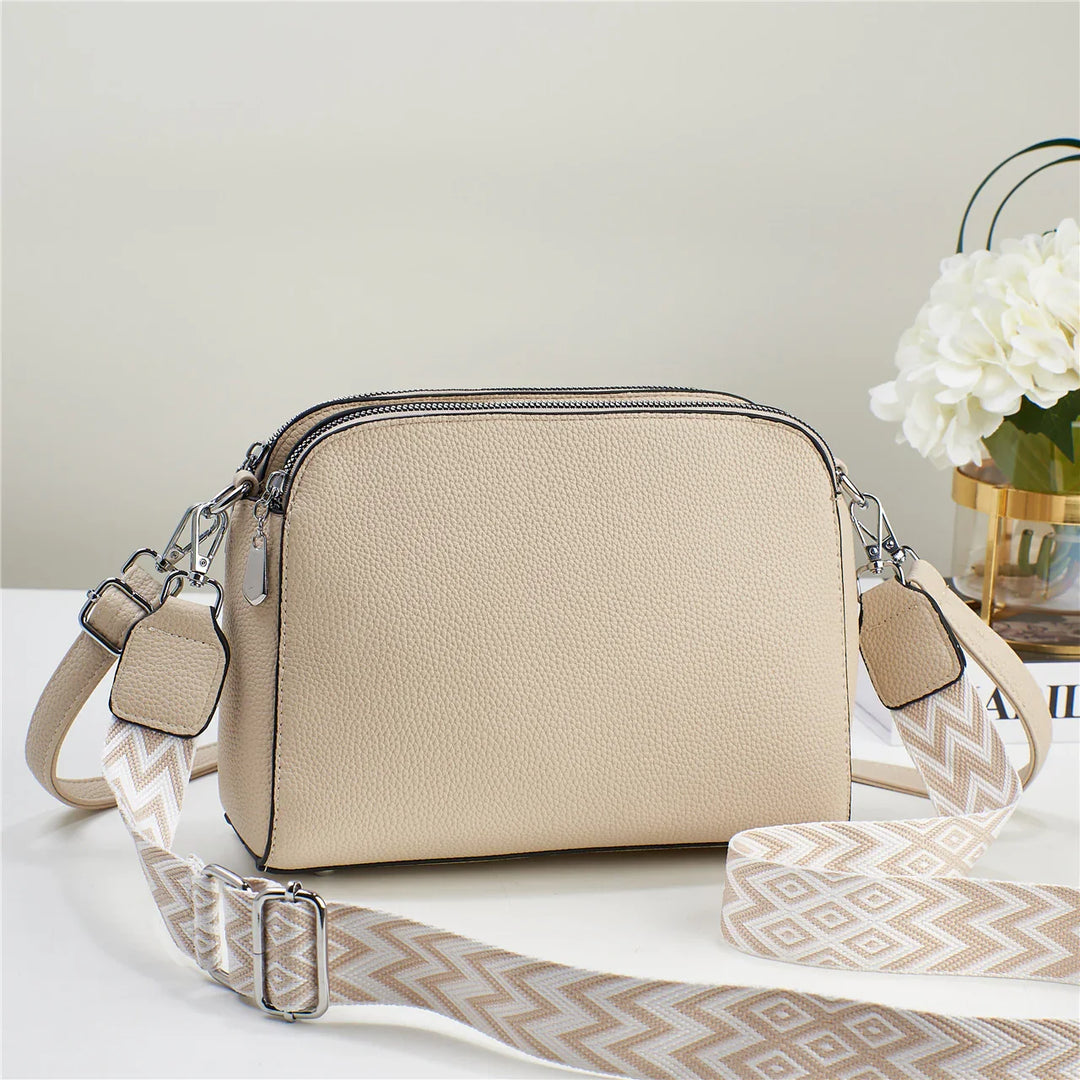 Beige textured leather crossbody bag with double zippers and patterned adjustable strap on white surface