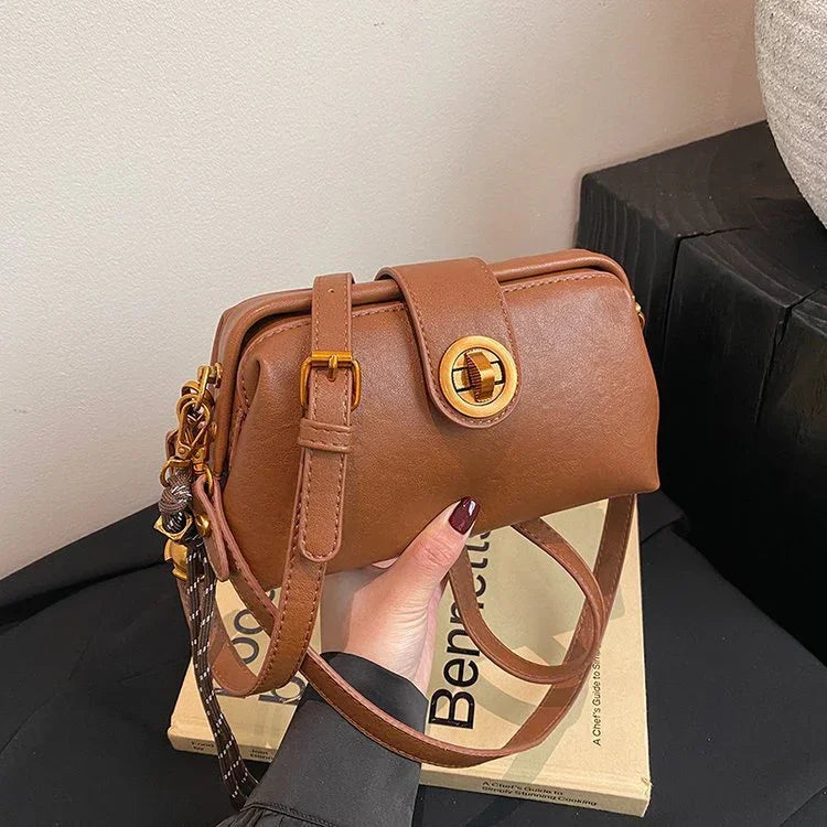 Hand holding a brown leather crossbody bag with gold buckle detailing on a book background