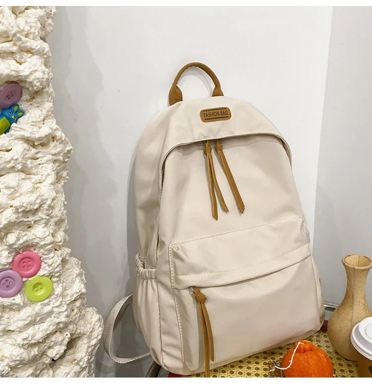 Beige fashion backpack with brown zipper pulls on a table next to a clay vase and textured wall with colorful buttons
