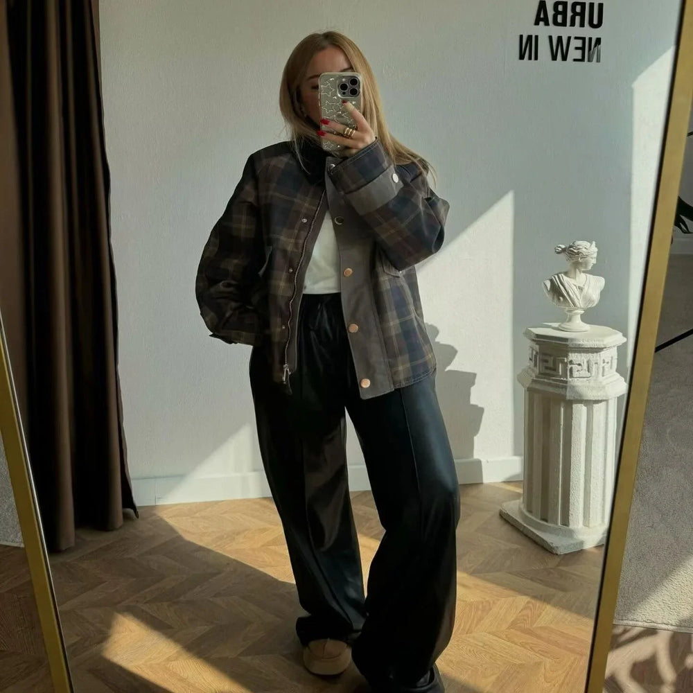 Woman taking mirror selfie wearing plaid jacket, white shirt, black wide pants, and slippers in sunlit room
