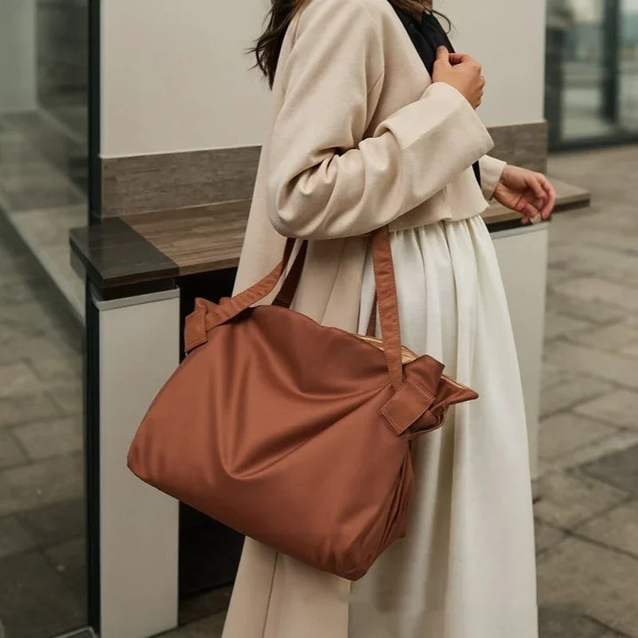 Woman wearing beige coat and white dress carrying a large brown leather tote bag outdoors