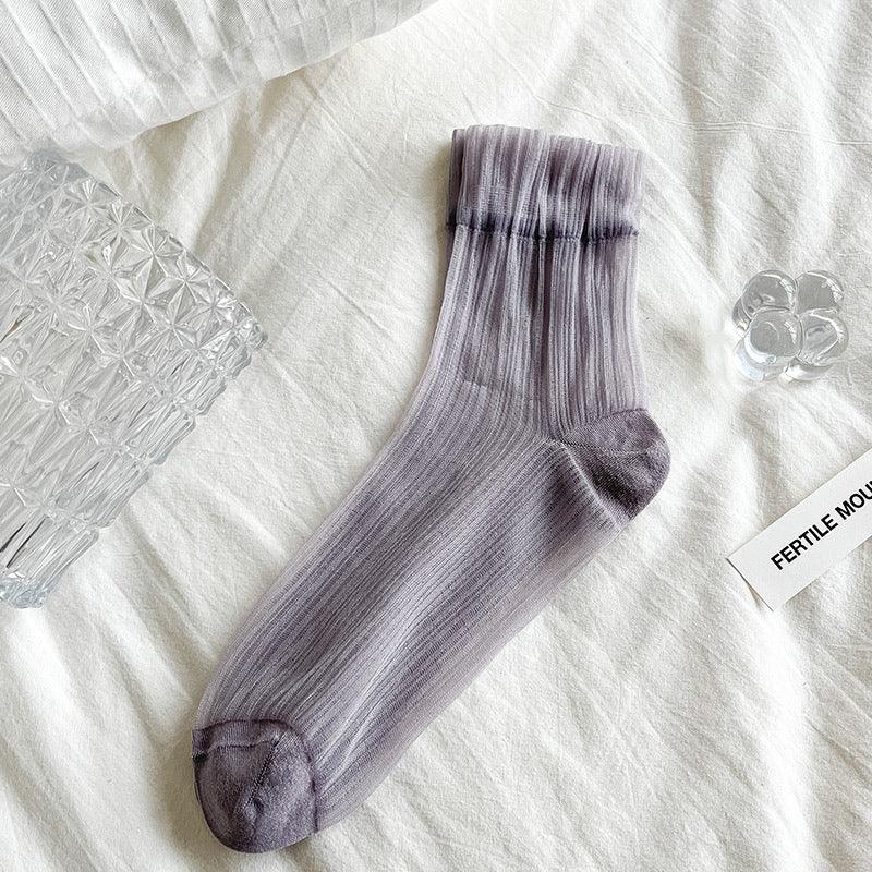 Single semi-transparent gray ribbed sock with darker toe and cuff laid on white textured fabric beside clear glass and white card