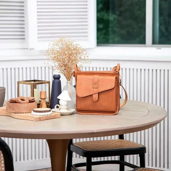 Tan leather crossbody bag on round table with dried flowers, cookies, and decorative items