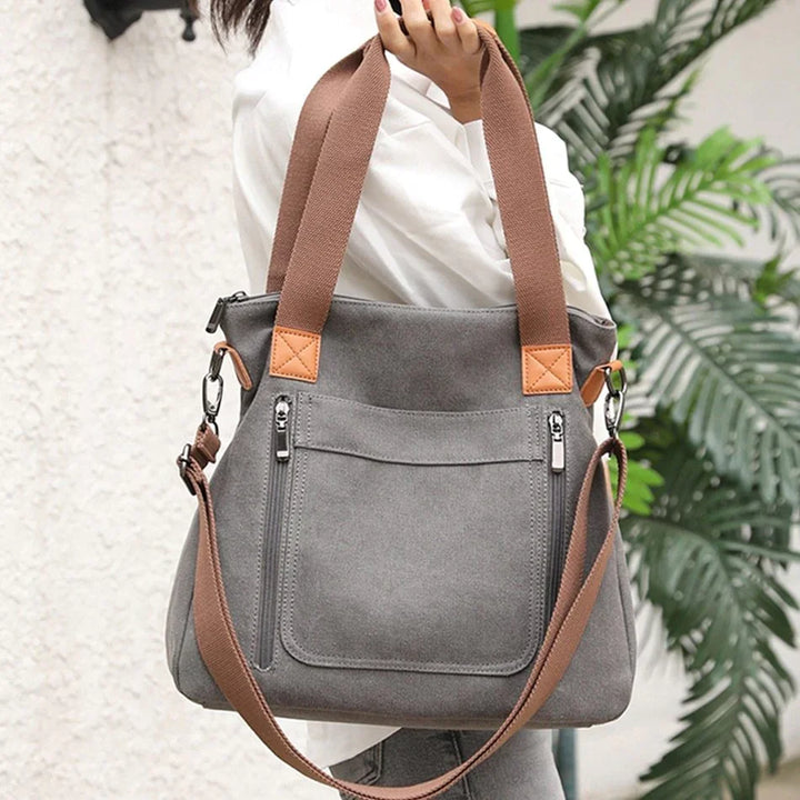 Gray suede tote bag with brown handles and adjustable strap held by person in white shirt near green plant