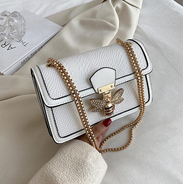 White textured leather handbag with gold chain strap and bee-shaped clasp, held by hand with red nail polish