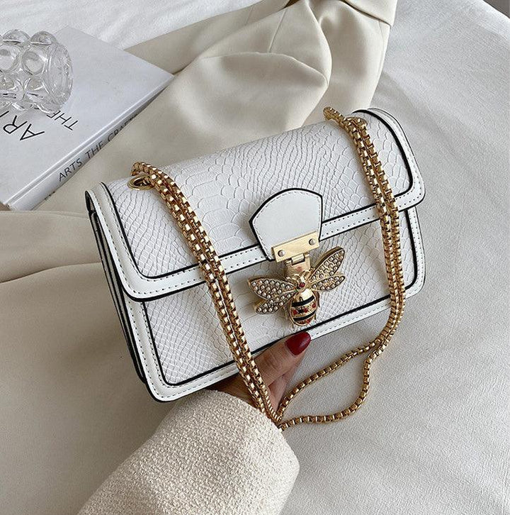White textured leather handbag with gold chain strap and bee-shaped clasp, held by hand with red nail polish