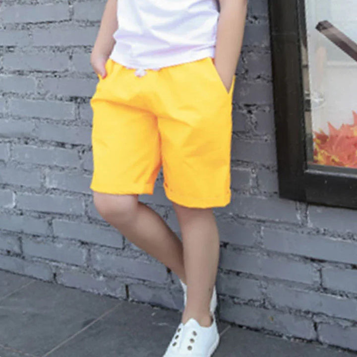 Child wearing bright yellow shorts and white sneakers standing against gray brick wall