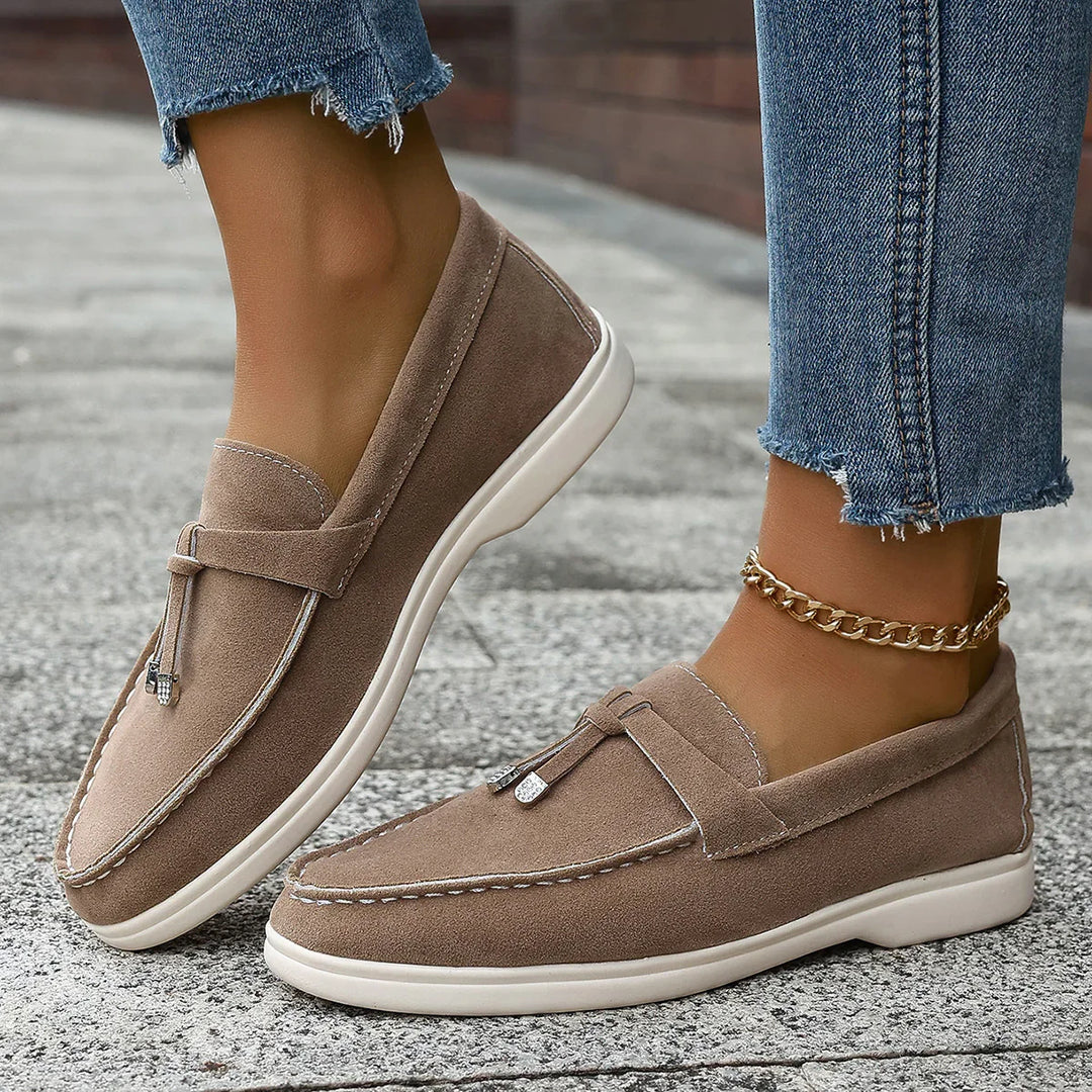 Brown suede loafers with tassels worn with frayed hem jeans and gold anklet on pavement