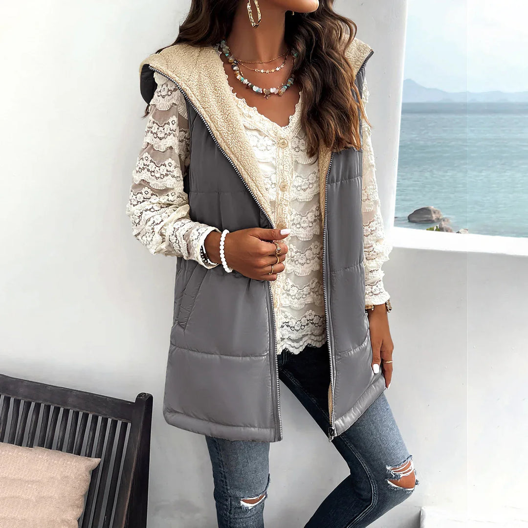 Woman wearing gray sherpa-lined hooded vest over white lace blouse and ripped jeans by seaside balcony