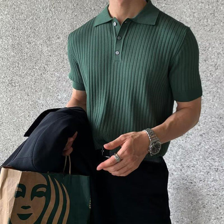 Man wearing green ribbed polo shirt holding black jacket and Starbucks paper bag against gray wall