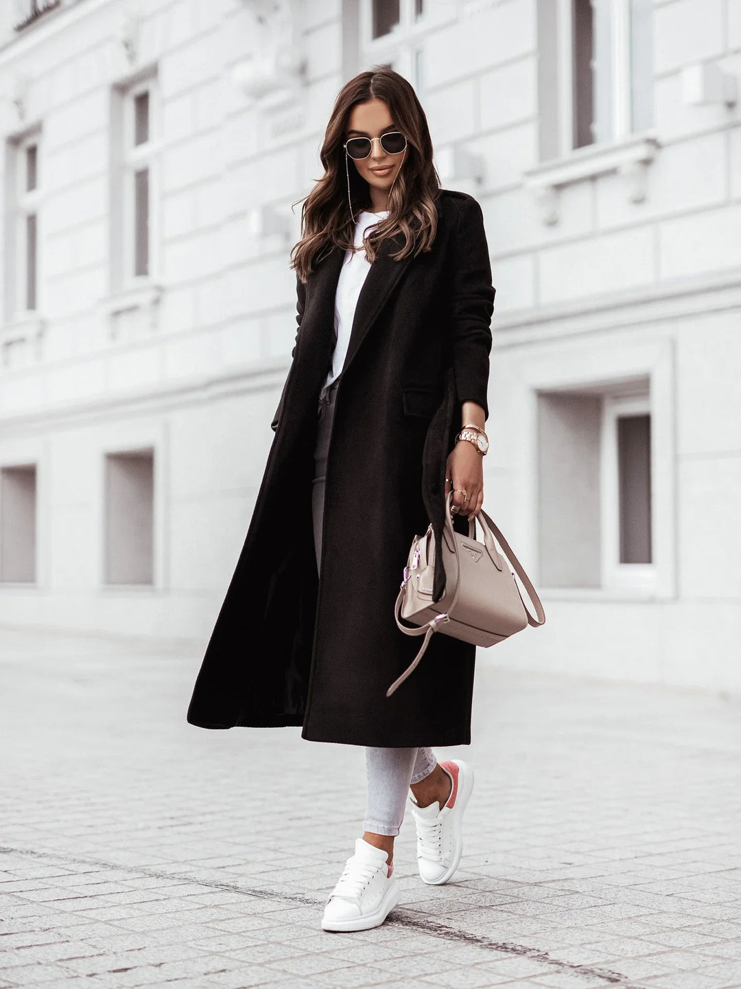 Woman in black long coat, white sneakers, sunglasses, and beige handbag walking on city street