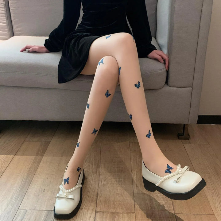 Person wearing beige tights with blue butterfly prints and white pearl-embellished shoes sitting on a gray sofa