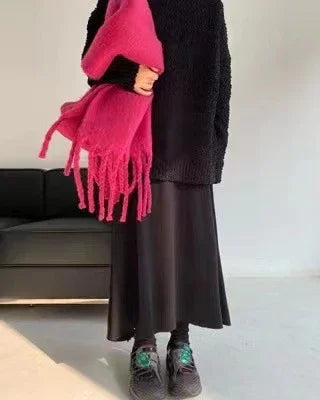 Person wearing a black sweater and skirt holding a bright pink fringed scarf, standing in a minimal room with a black sofa