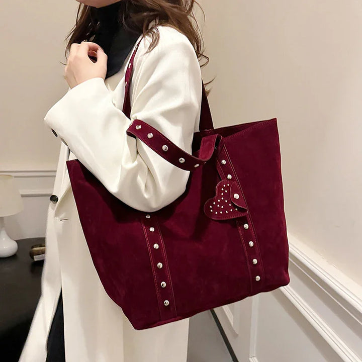 Woman in white coat carrying large burgundy suede tote bag with studded straps and heart charm