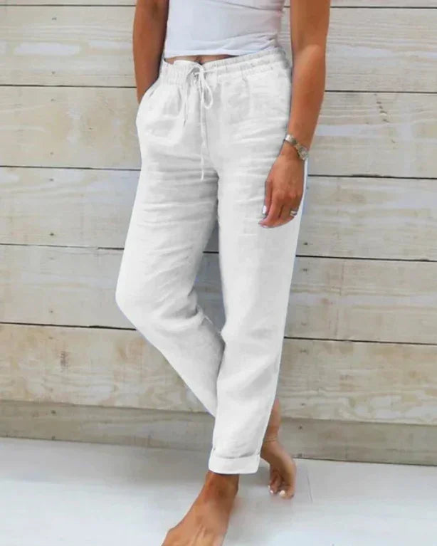 Woman wearing white drawstring linen pants and a white top standing barefoot against a wood plank wall