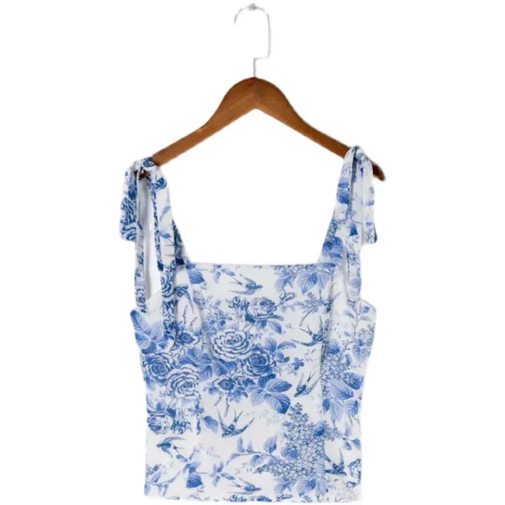 White camisole top with blue floral and bird print, tied shoulder straps, hanging on wooden hanger