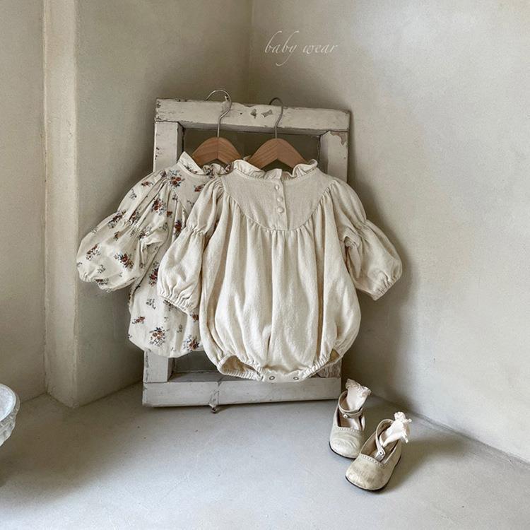 Vintage-style baby rompers and floral dress hanging on rustic wooden frame with beige baby shoes on floor