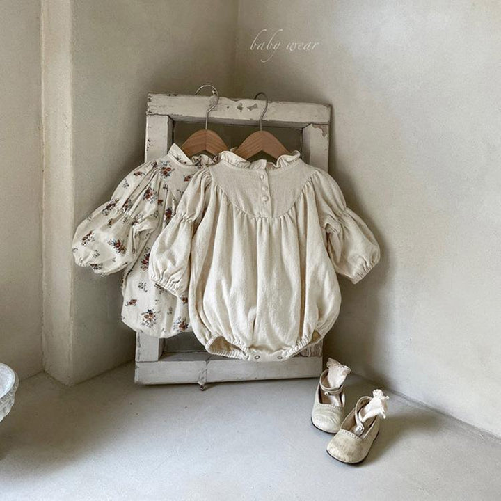 Vintage-style baby rompers and floral dress hanging on rustic wooden frame with beige baby shoes on floor