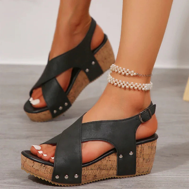 Women's black cork wedge sandals with crisscross straps and silver studs, paired with pearl ankle bracelets