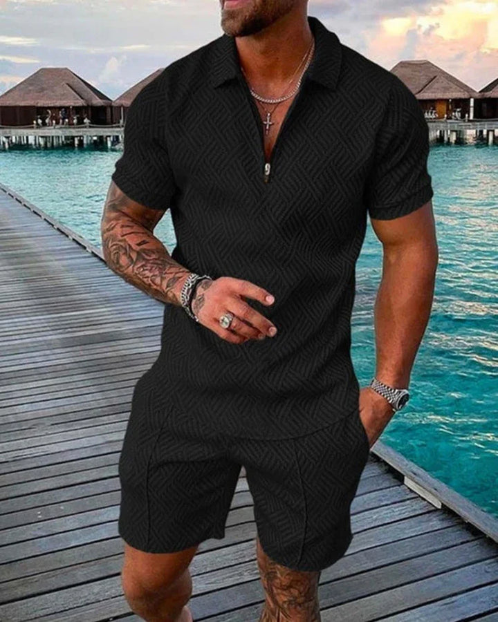 Muscular man wearing black patterned short-sleeve shirt and shorts, standing on wooden dock by blue ocean with overwater bungalows