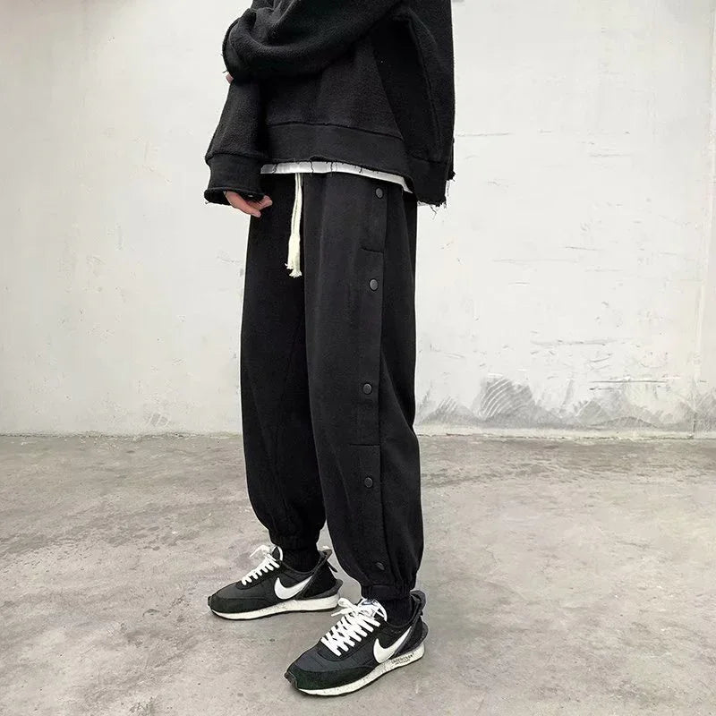 Person wearing black jogger pants with side buttons, black Nike sneakers, and a black oversized sweatshirt in a minimal indoor setting