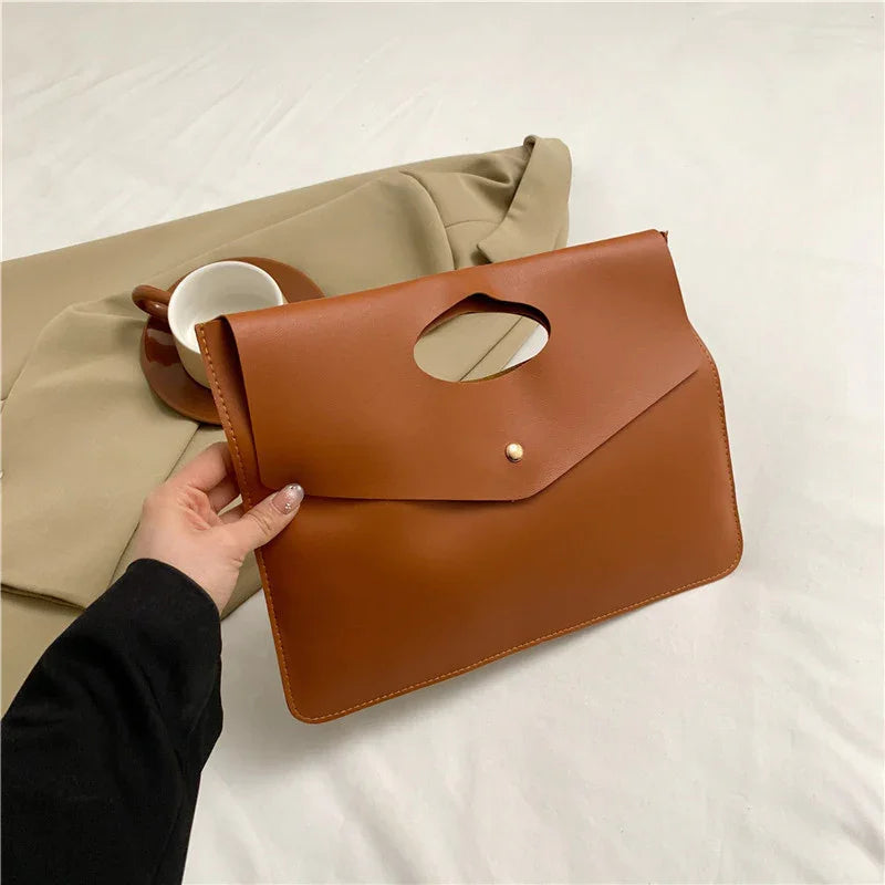 Hand holding brown leather envelope clutch bag over beige fabric with brown coffee cup and saucer