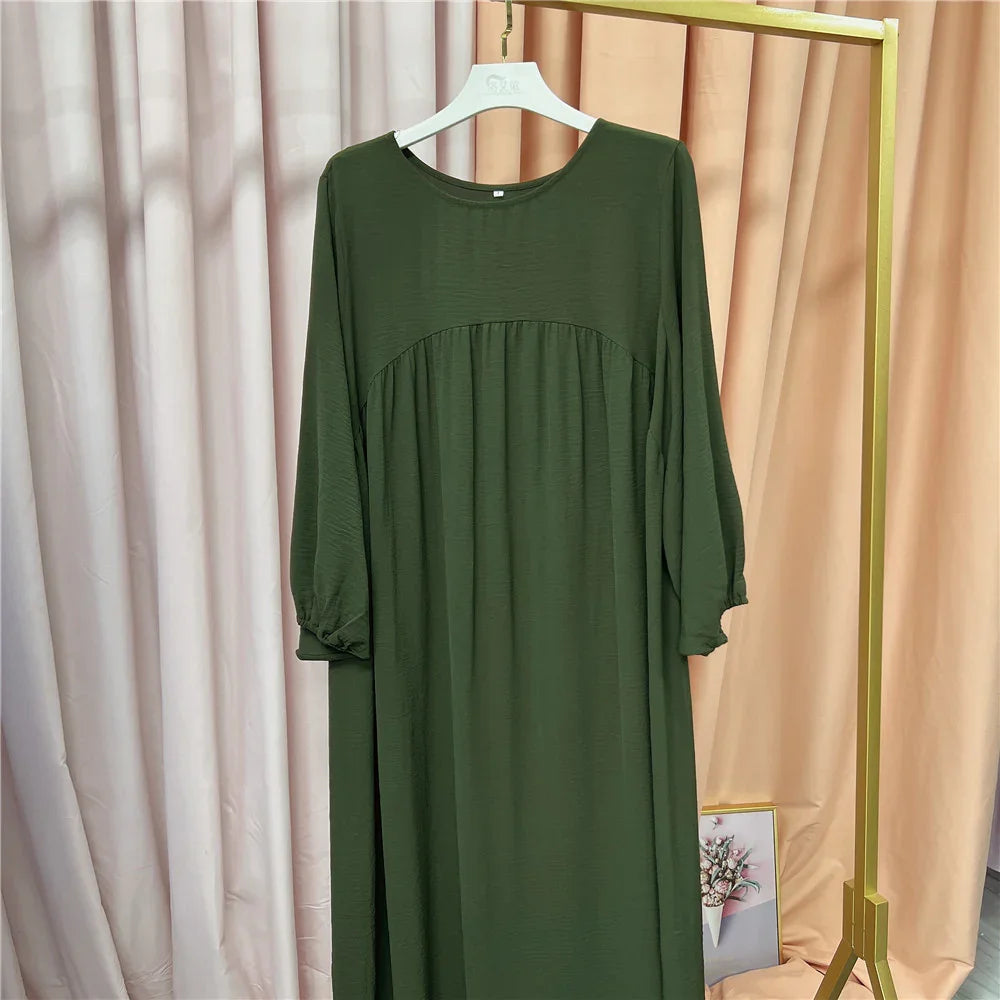 Long olive green dress with three-quarter sleeves hanging on white hanger against beige and pink curtains