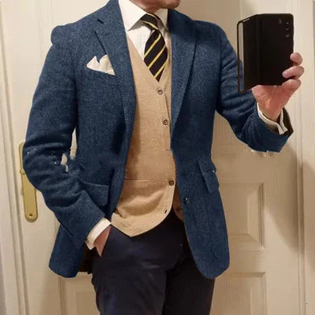 Man wearing blue blazer, beige cardigan, striped tie, navy pants, white pocket square taking mirror selfie indoors