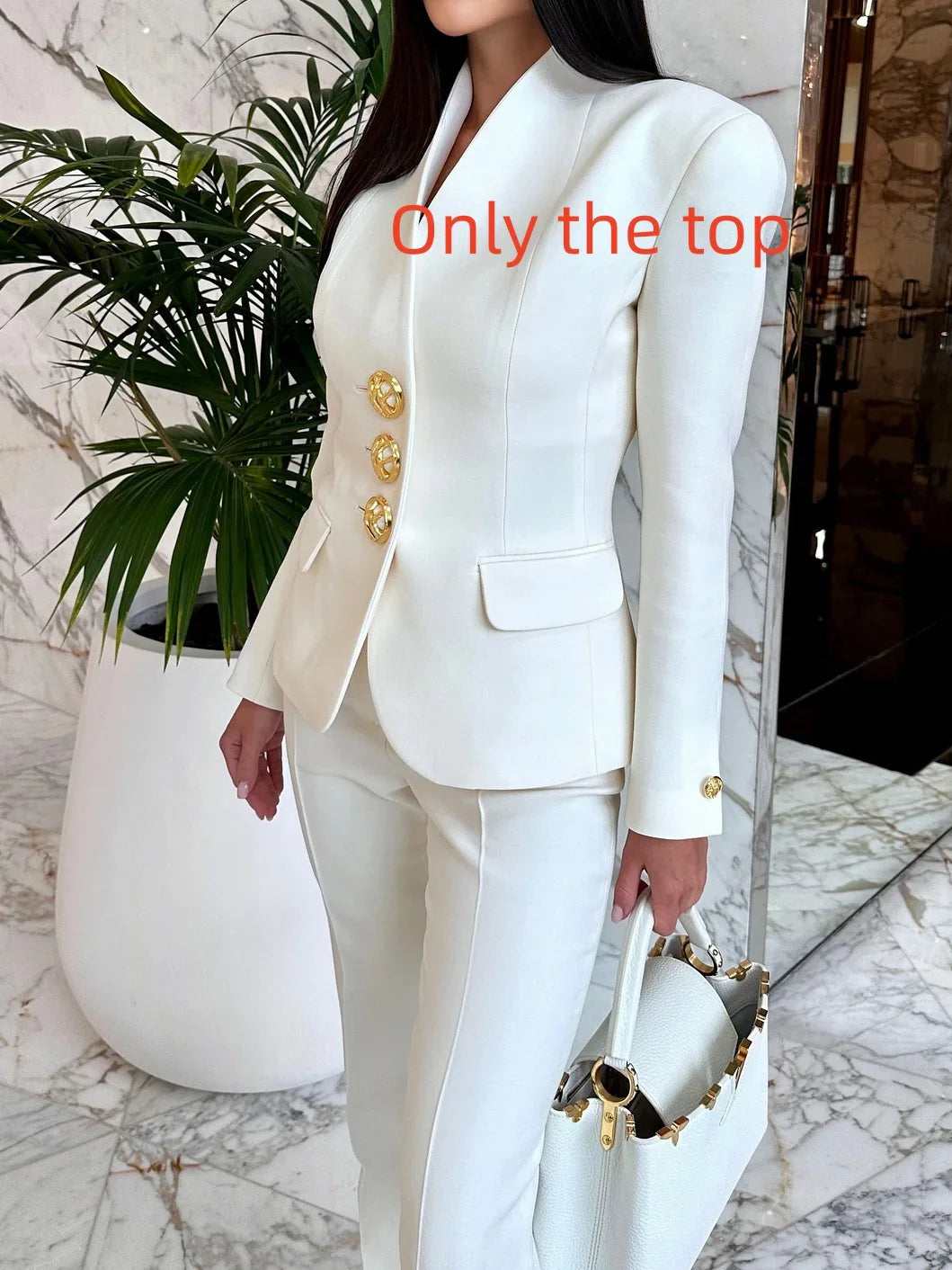 Woman wearing white tailored blazer with gold buttons and matching pants holding white handbag indoors