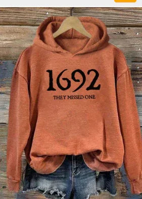 Rust-colored hoodie with black text '1692 THEY MISSED ONE' displayed on wooden hanger over jean shorts