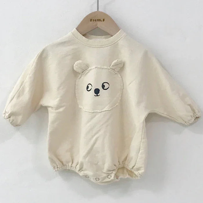 Cream-colored baby romper with bear face and ears design, displayed on wooden hanger against white wall