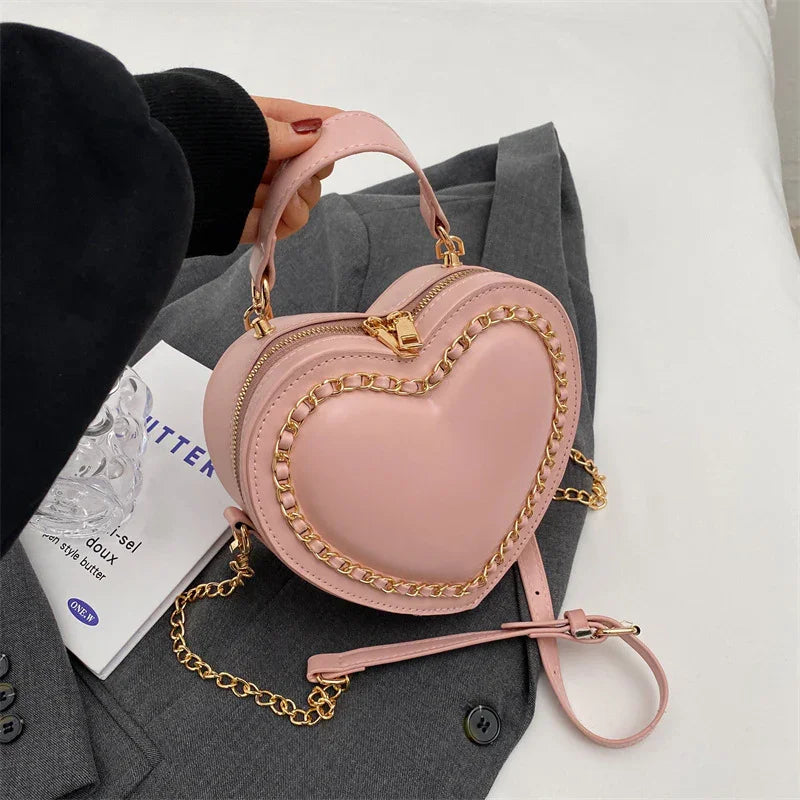 Pink heart-shaped handbag with gold chain detail, held by hand over gray jacket and white book
