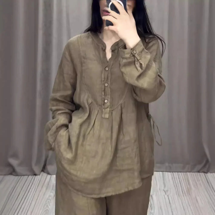 Woman wearing loose brown linen blouse with buttons, standing in front of gray curtain background