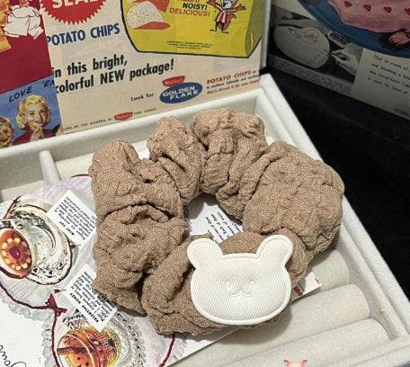 Beige fabric scrunchie with white bear face patch in a jewelry display box with retro chip bag background