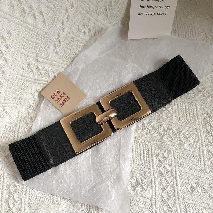 Black elastic belt with gold rectangular buckle on white textured fabric with 'Que Sera Sera' card
