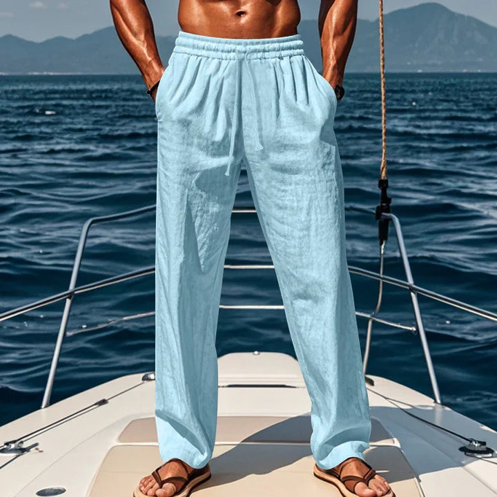 Muscular man wearing light blue linen pants and brown sandals on yacht deck with ocean background