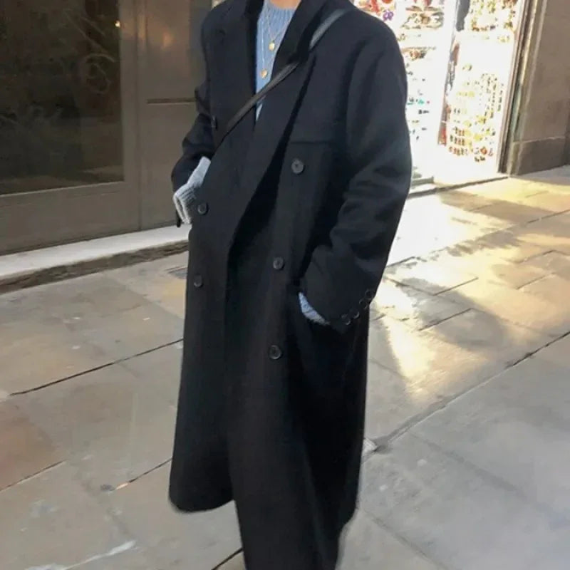 Person wearing long black double-breasted coat over a blue sweater on city sidewalk at night