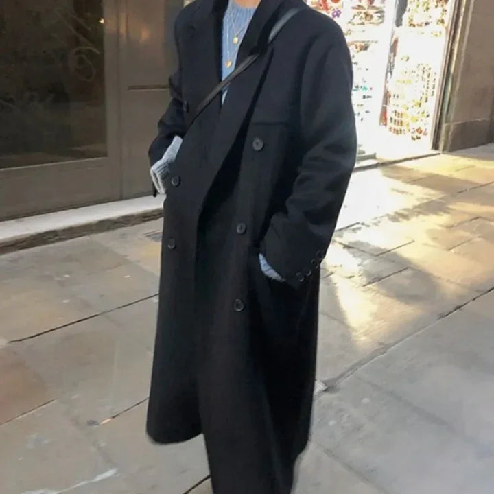 Person wearing long black double-breasted coat over a blue sweater on city sidewalk at night