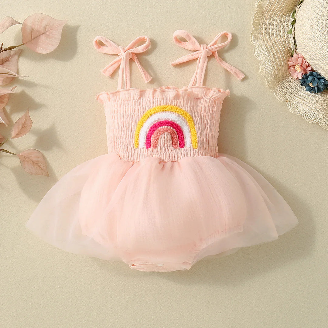 Pink baby romper with tulle skirt and rainbow embroidery, displayed with straw hat and dried leaves