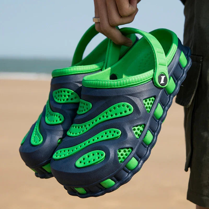 Hand holding navy blue and bright green ventilated slip-on sandals on a sandy beach background