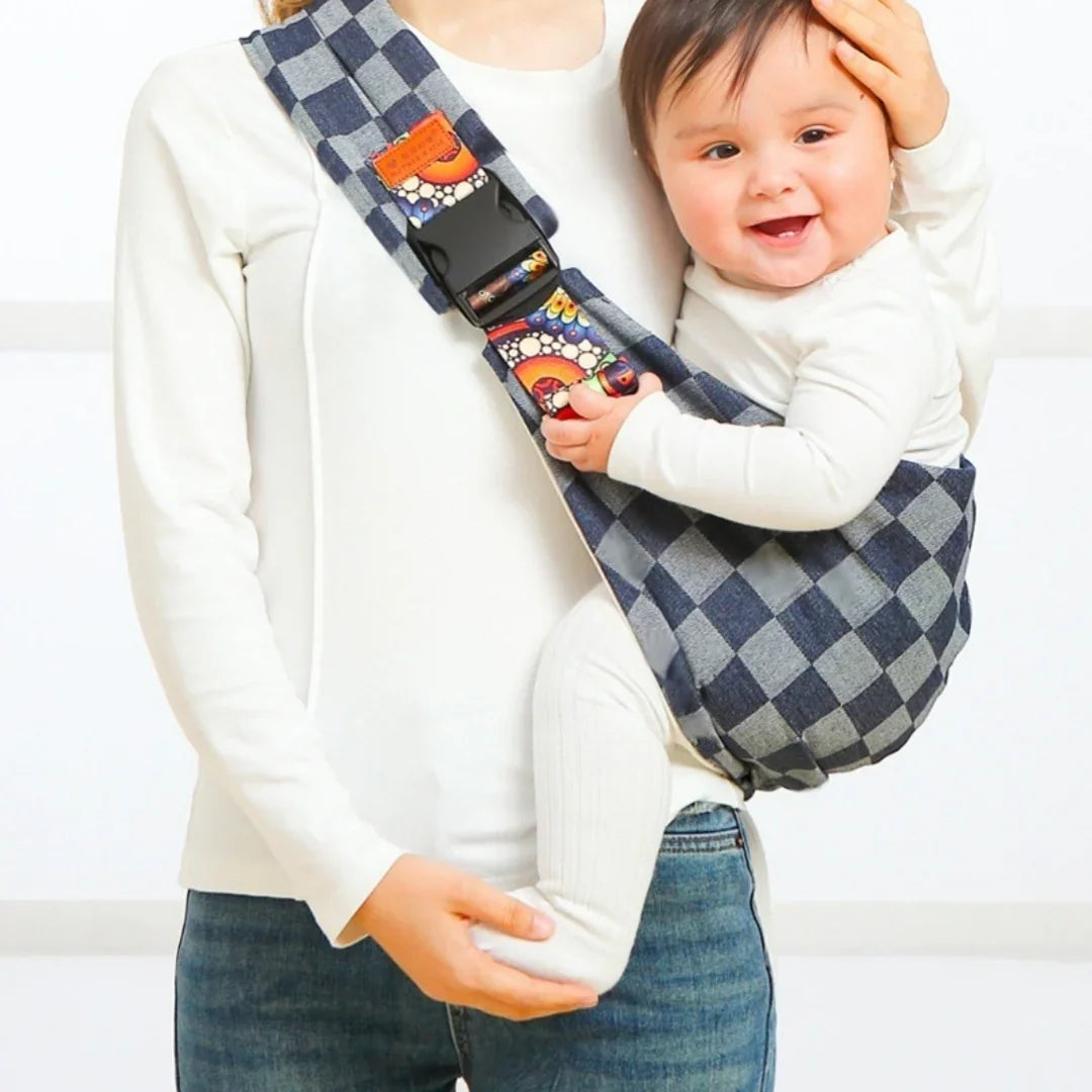 Smiling baby in checkered blue sling carrier worn by person in white top and jeans