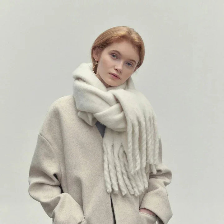 Young woman with red hair wearing oversized beige coat and thick white scarf against plain background