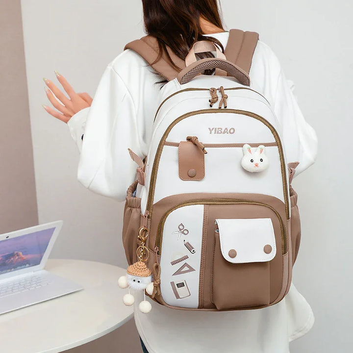 White and brown YIBAO backpack with school supplies design, bunny keychain, worn by person in white shirt