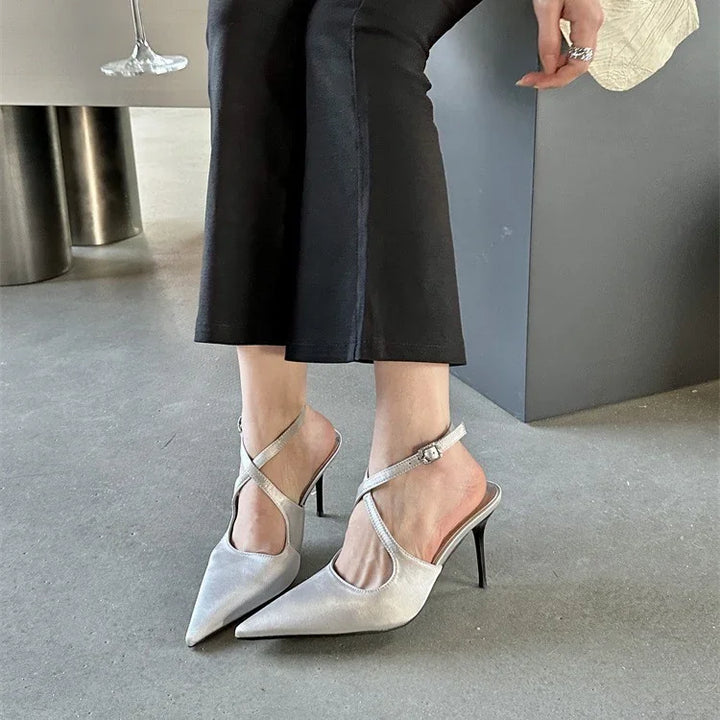 Woman wearing silver pointed toe stiletto heels with criss-cross ankle straps and black cropped pants indoors
