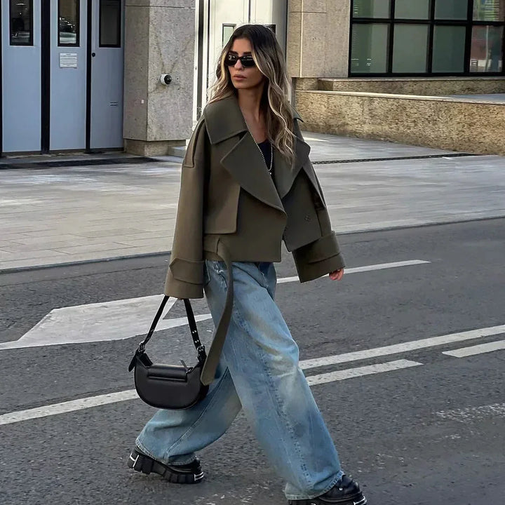 Woman walking on street wearing oversized olive green jacket, baggy light blue jeans, black chunky shoes, and sunglasses