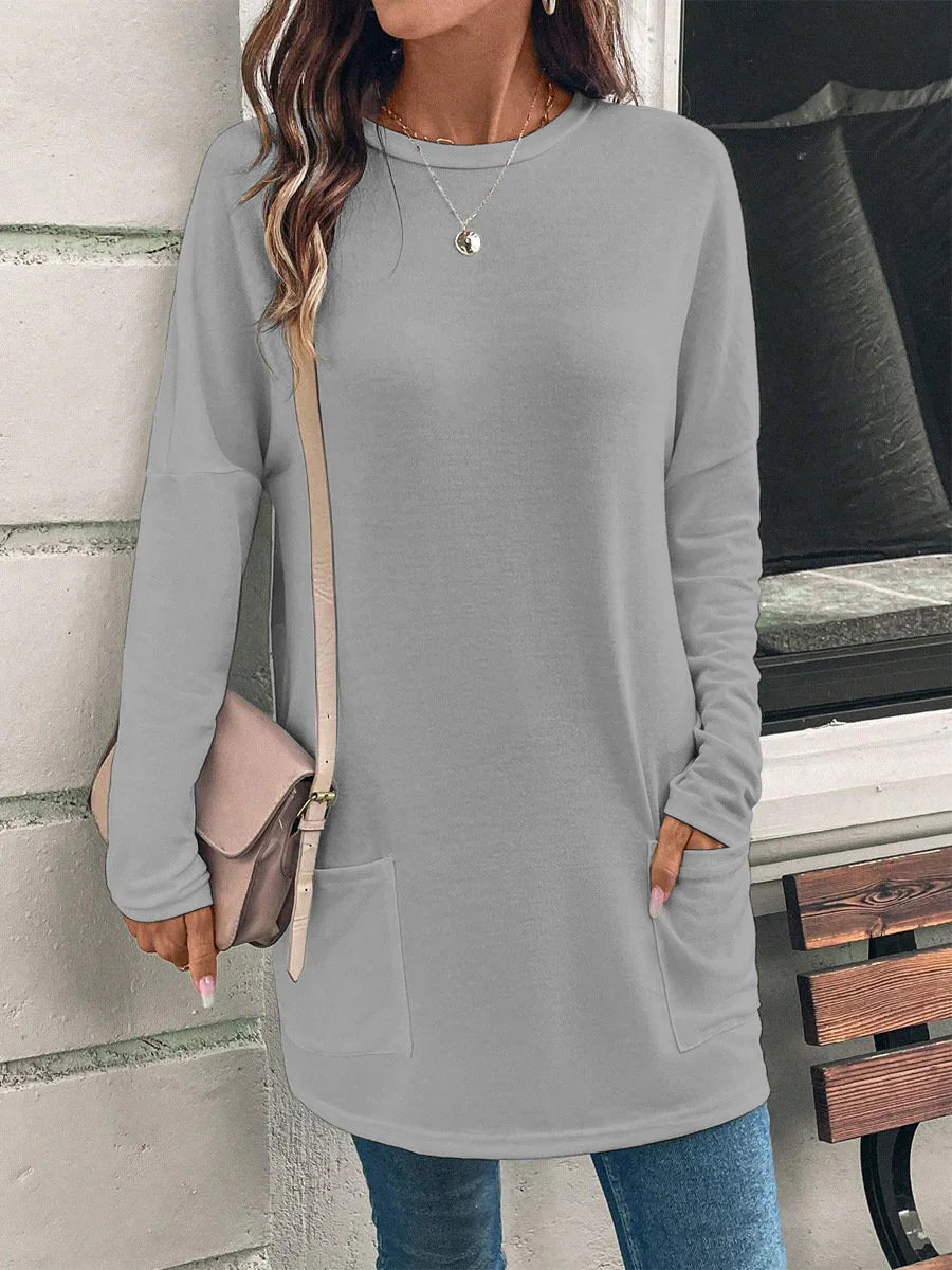 Woman wearing light gray long sleeve tunic sweater with pockets, blue jeans, and beige shoulder bag