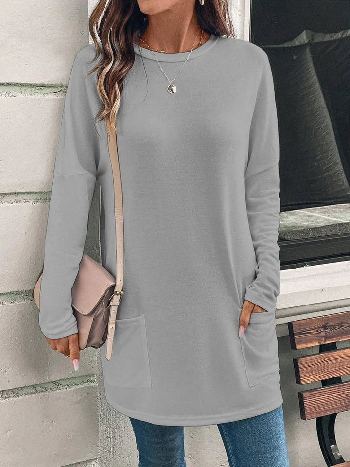 Woman wearing light gray long sleeve tunic sweater with pockets, blue jeans, and beige shoulder bag