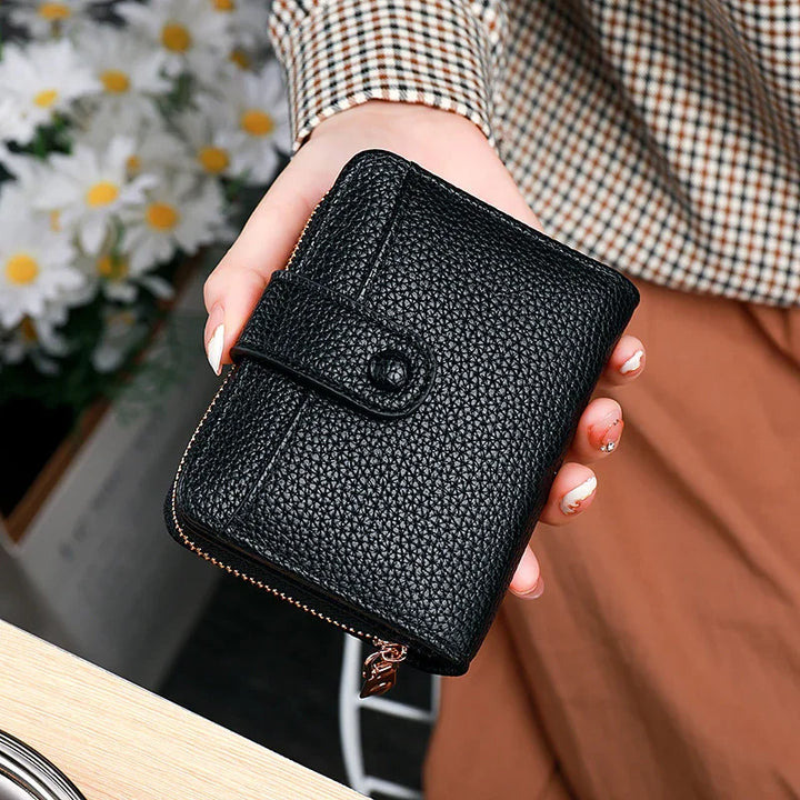 Hand holding black textured leather wallet with zipper and button clasp, checkered shirt and daisies background